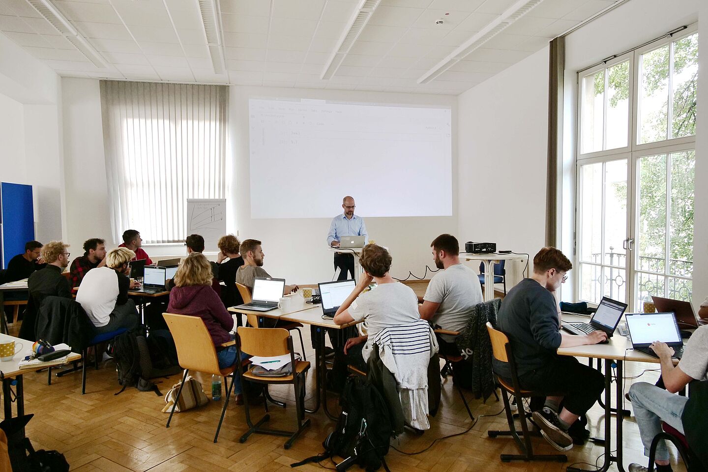 HTWK-Professor Gerold Bausch steht vorn, via Beamer wird ein Bild an die Wand projiziert. Die Studierenden sitzen an Tischen und hören zu.