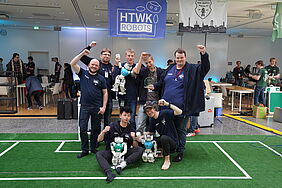 Menschen mit humanoiden Robotern auf kleinem Fußballfeld indoor