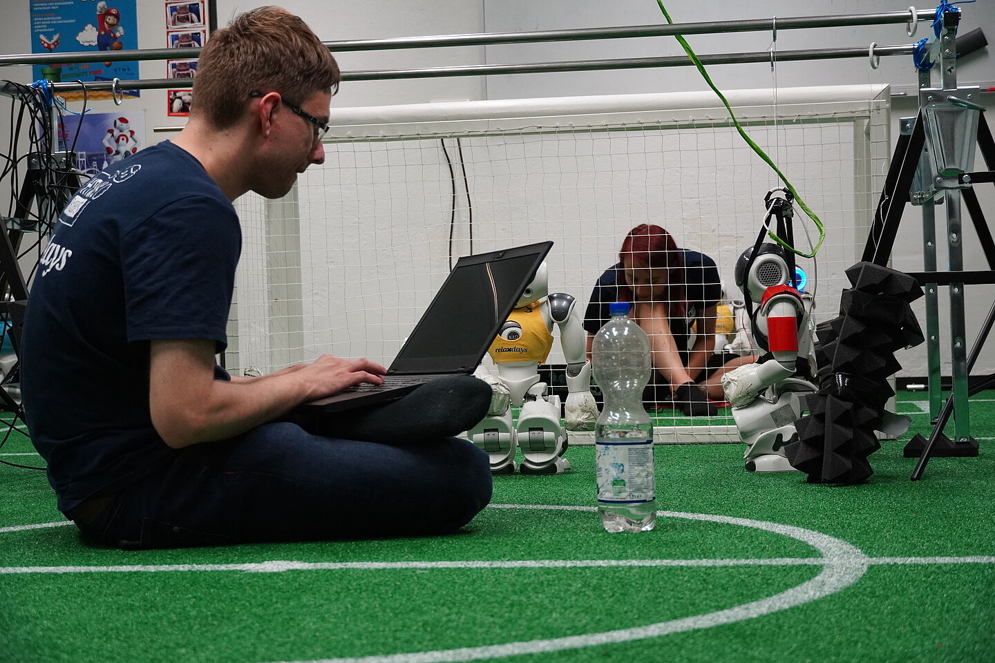 Teammitglieder Tobias Jagla und Lea Kunz (hinten im Bild) bei den Vorbereitungen. Mensch sitzt auf grünem Teppich mit Robotern und Fußballtor