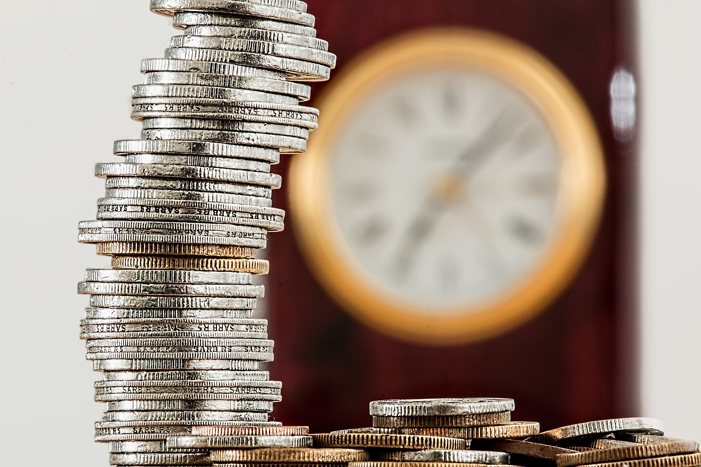 Zeit ist Geld Zeit ist Geld: Zu sehen sind ein hoher Münzstapel und dahinter verschwommen eine Uhr.