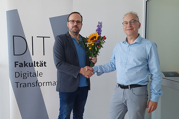 Prof. Schemmert (rechts) schüttelt Prof. Crönertz (links) die Hand, der Blumen in der anderen Hand hält. Rollup mit FDIT-Logo ist im Hintergrund.